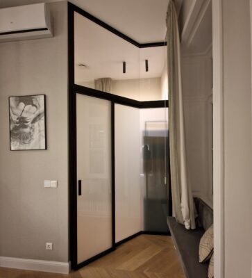 Corner of a modern room with a glass-front wardrobe, beige walls, and a bench with patterned cushions by the window.