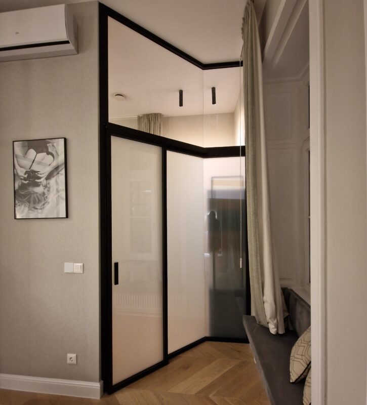 Corner of a modern room with a glass-front wardrobe, beige walls, and a bench with patterned cushions by the window.