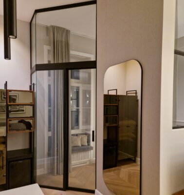 Showroom interior with large glass-front wardrobes in black frames, reflecting a window and light curtains, and light wood flooring.