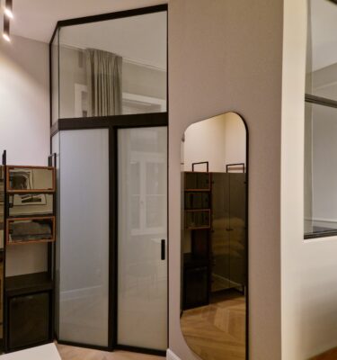 Showroom scene with tall glass-front wardrobes in black frames, beige walls, and a curved-edge mirror on the right; wooden floor visible.